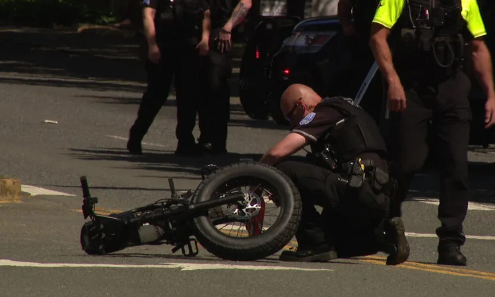 Officer kneels beside a toppled motorcycle on a city street as other officers stand nearby.