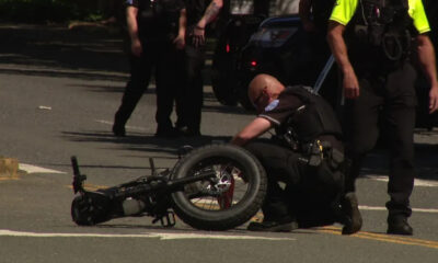 Officer kneels beside a toppled motorcycle on a city street as other officers stand nearby.