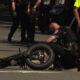 Officer kneels beside a toppled motorcycle on a city street as other officers stand nearby.