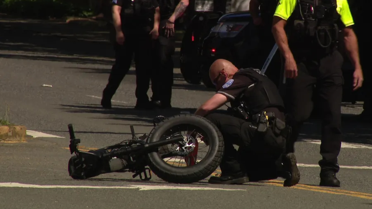 Officer kneels beside a toppled motorcycle on a city street as other officers stand nearby.