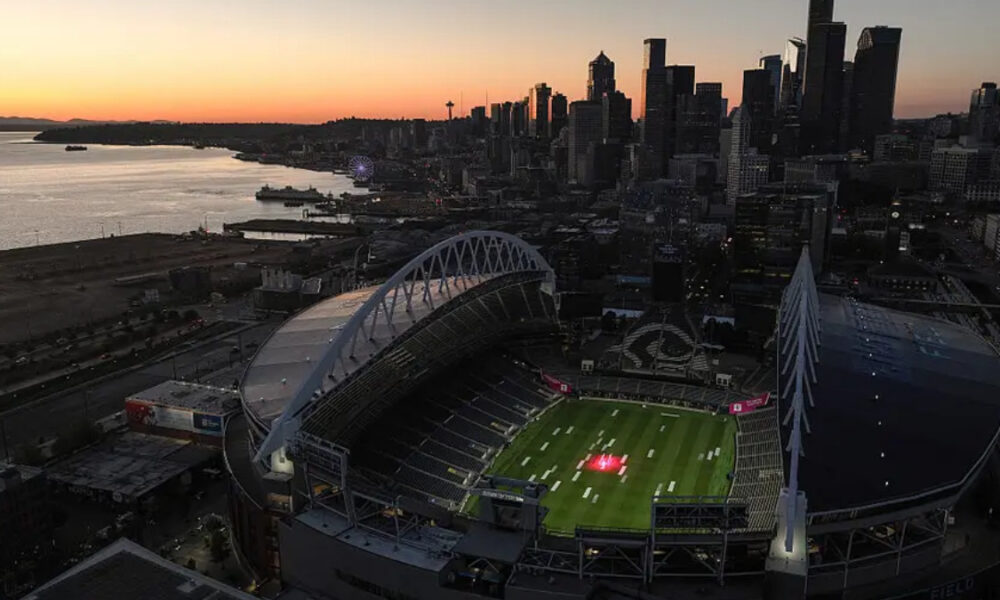 Aerial view of a large football stadium at sunset, with a green field and a city skyline along the waterfront in the background.