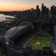 Aerial view of a large football stadium at sunset, with a green field and a city skyline along the waterfront in the background.
