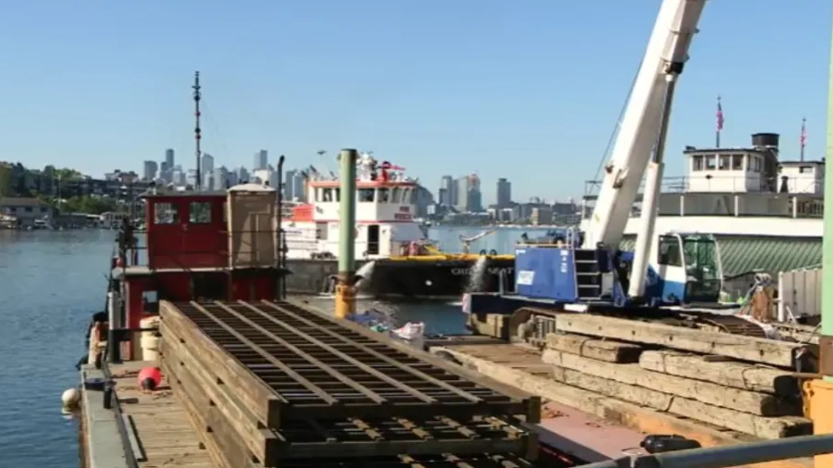 Dockside construction area with stacked steel beams, a blue crane, and boats on calm water; city skyline in the background.