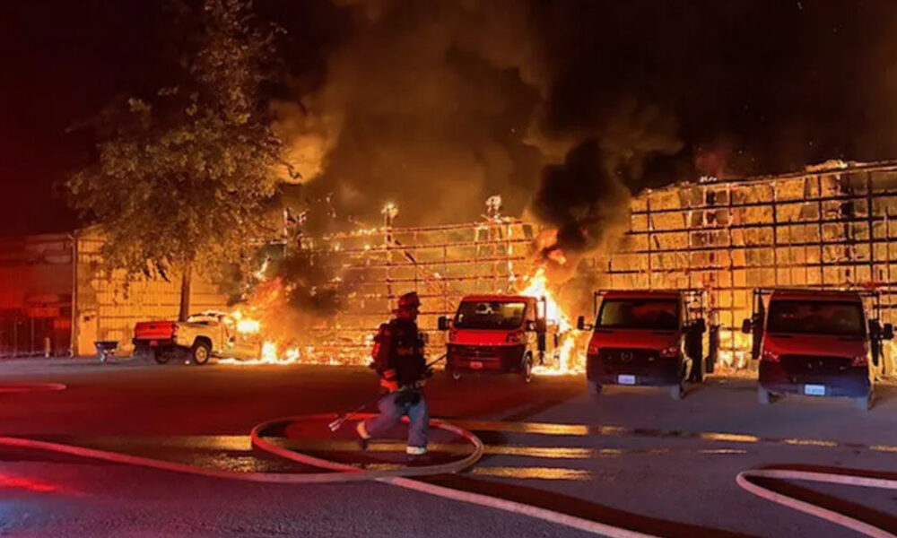 Nighttime scene of a firefighter in turnout gear directing a hose toward a burning warehouse as flames engulf the structure and thick black smoke rises, with several red vans and a pickup truck parked in front.