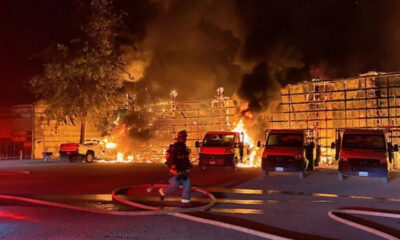 Nighttime scene of a firefighter in turnout gear directing a hose toward a burning warehouse as flames engulf the structure and thick black smoke rises, with several red vans and a pickup truck parked in front.