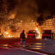 Nighttime scene of a firefighter in turnout gear directing a hose toward a burning warehouse as flames engulf the structure and thick black smoke rises, with several red vans and a pickup truck parked in front.