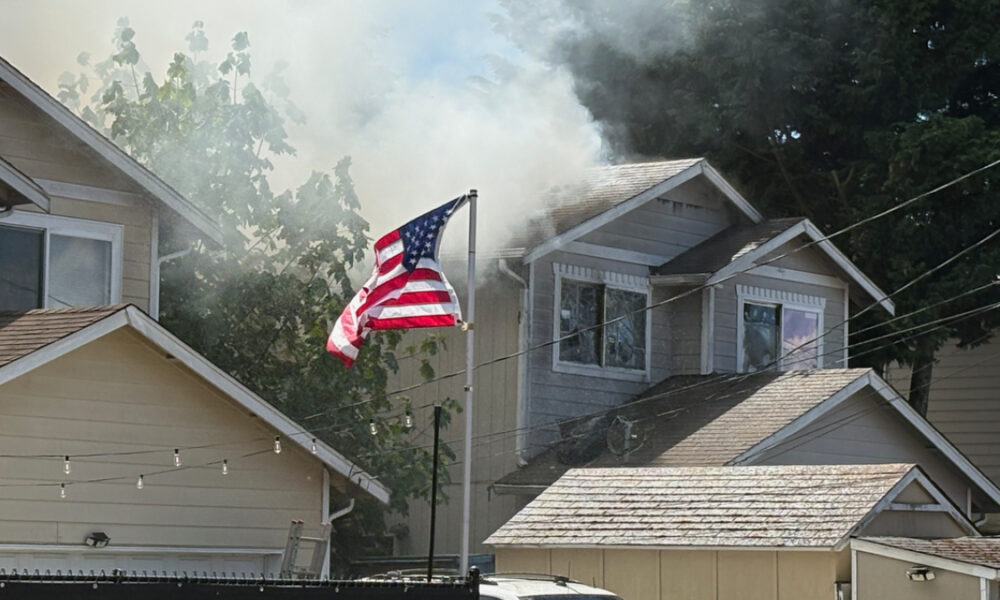 American flag flaps in front of a two-story house as smoke rises from the roof.