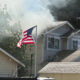 American flag flaps in front of a two-story house as smoke rises from the roof.