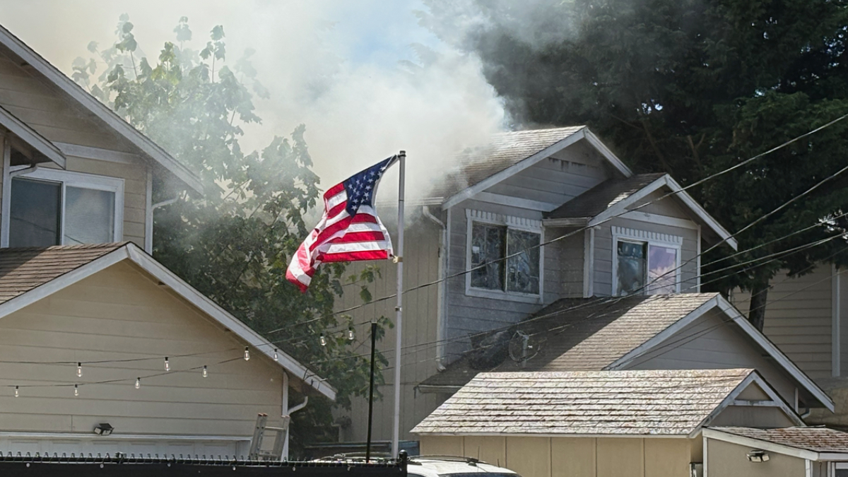 American flag flaps in front of a two-story house as smoke rises from the roof.
