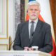 Man in a gray suit and tie speaks at a podium with microphones, flags in the background.