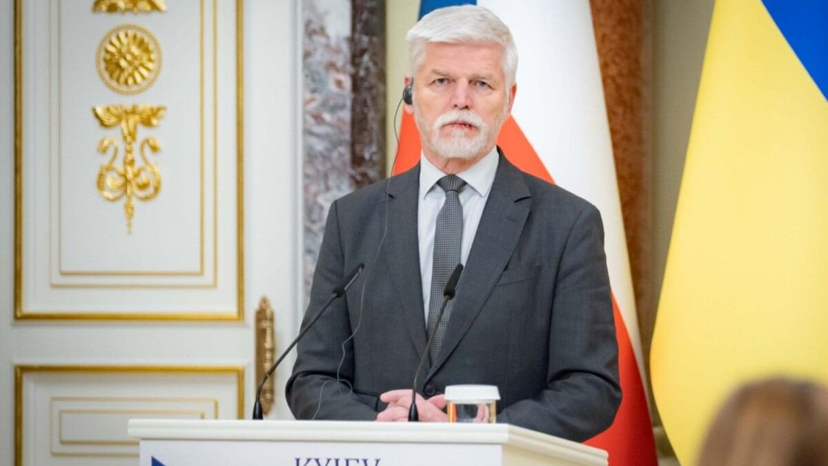 Man in a gray suit and tie speaks at a podium with microphones, flags in the background.