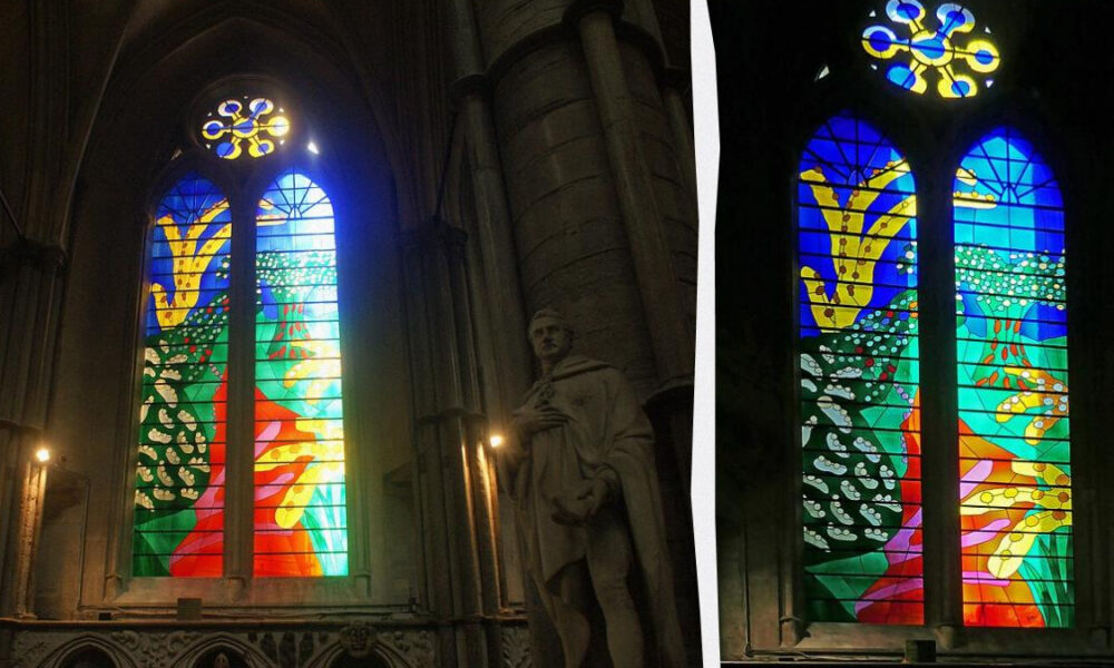 Stained glass church window with vibrant abstract landscape in blue, green, red, and yellow hues; stone arches nearby and a statue in foreground (left).