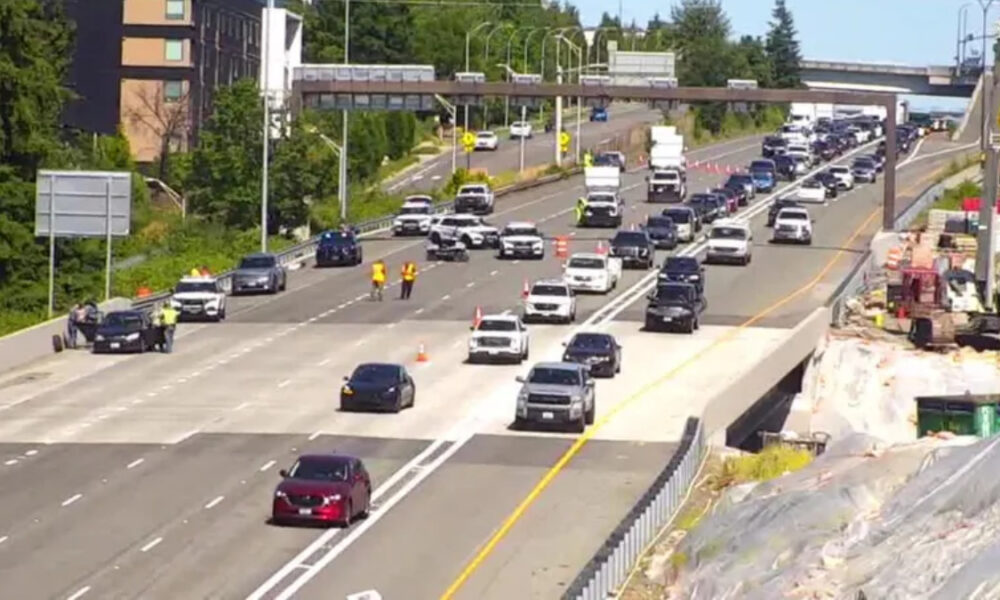 Busy multi-lane highway with construction on the right, traffic moving slowly and orange cones marking a lane closure behind a guardrail area nearby