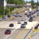 Busy multi-lane highway with construction on the right, traffic moving slowly and orange cones marking a lane closure behind a guardrail area nearby
