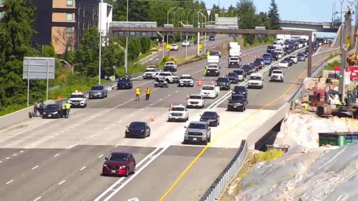 Busy multi-lane highway with construction on the right, traffic moving slowly and orange cones marking a lane closure behind a guardrail area nearby