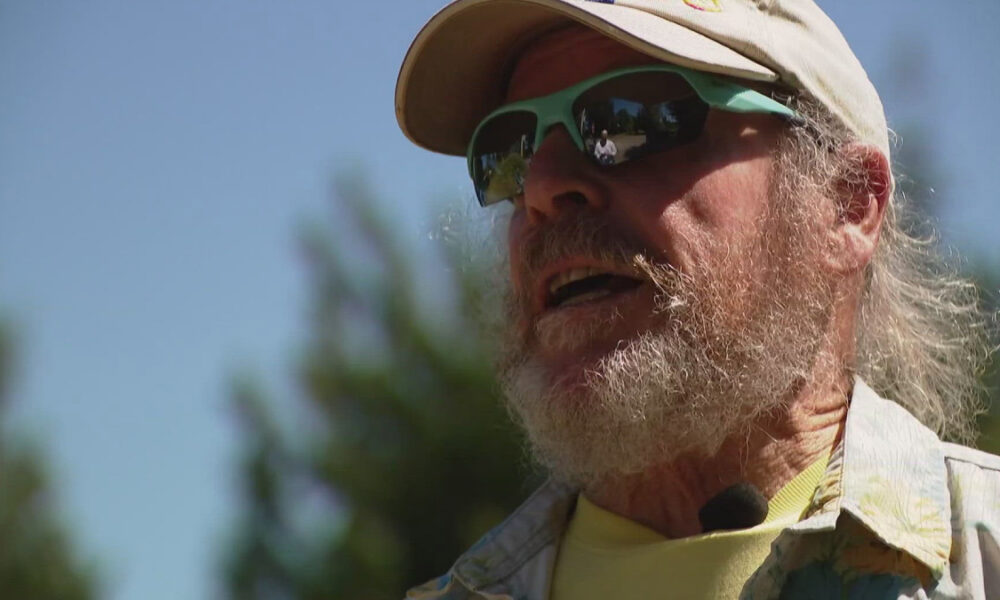 Older man with a gray beard wearing a beige cap and green sunglasses outdoors, speaking into a clip-on mic.