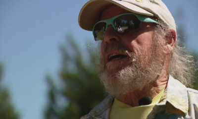 Older man with a gray beard wearing a beige cap and green sunglasses outdoors, speaking into a clip-on mic.