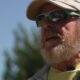 Older man with a gray beard wearing a beige cap and green sunglasses outdoors, speaking into a clip-on mic.