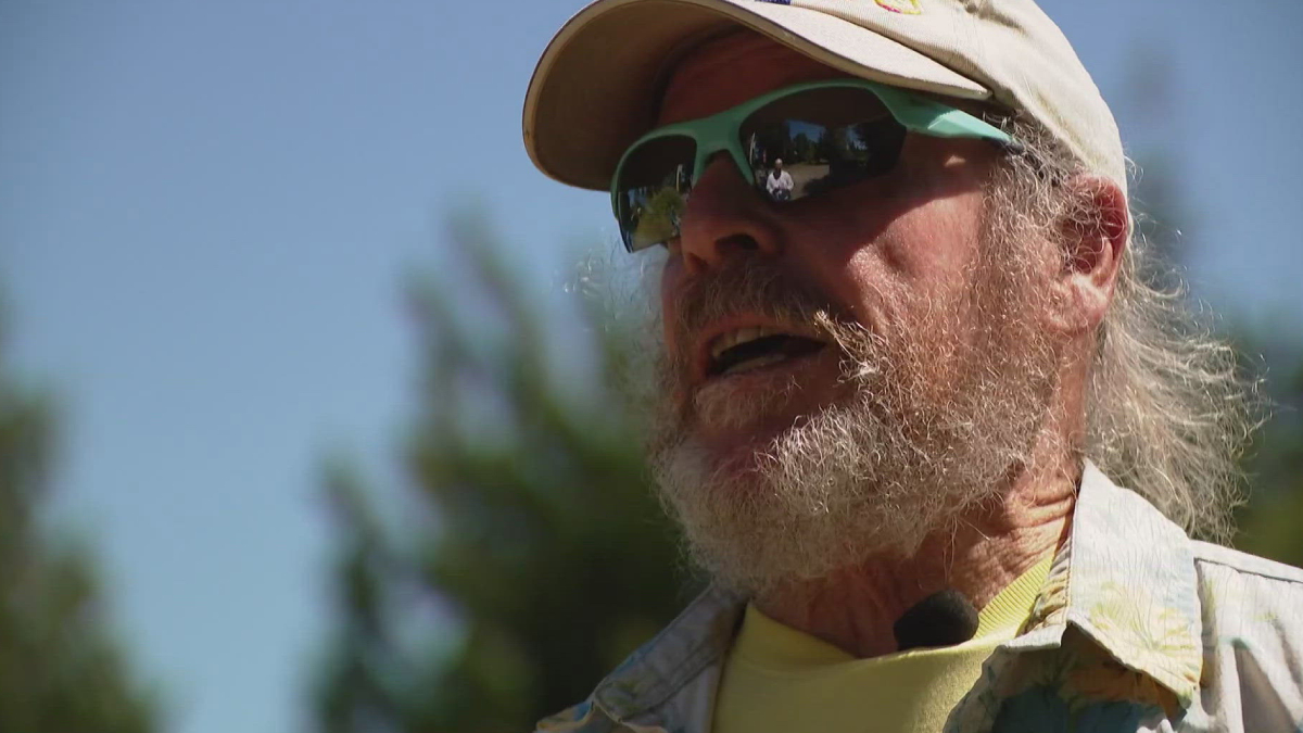 Older man with a gray beard wearing a beige cap and green sunglasses outdoors, speaking into a clip-on mic.