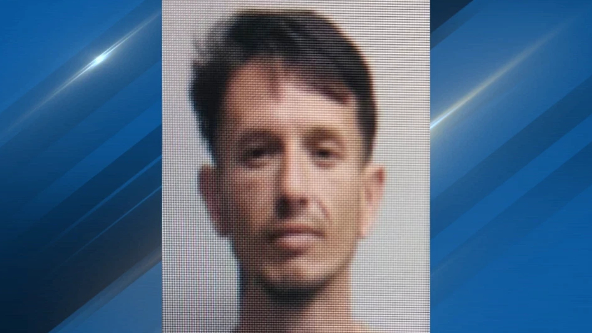 Mugshot-style portrait of a man with short dark hair and a neutral expression, gray backdrop behind him.