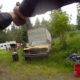 First-person view: two people shaking hands while a pistol is pointed toward the camera in a grassy junkyard with an old yellow car in the background.
