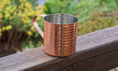 Hammered copper mug with a curved handle sits on a weathered wooden railing, outdoors with greenery in the background.