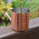Hammered copper mug with a curved handle sits on a weathered wooden railing, outdoors with greenery in the background.