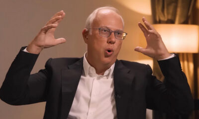 Man in a suit and glasses speaks animatedly, hands raised, during an interview or discussion set.