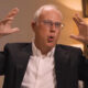 Man in a suit and glasses speaks animatedly, hands raised, during an interview or discussion set.