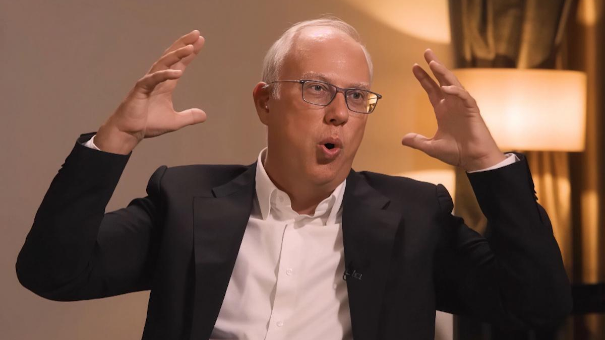 Man in a suit and glasses speaks animatedly, hands raised, during an interview or discussion set.