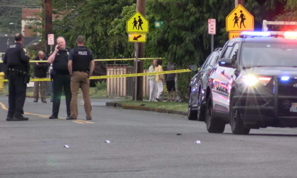 Police officers stand behind yellow crime-scene tape on a street, with flashing police car lights and bystanders in the background near a crosswalk sign.