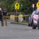 Police officers stand behind yellow crime-scene tape on a street, with flashing police car lights and bystanders in the background near a crosswalk sign.