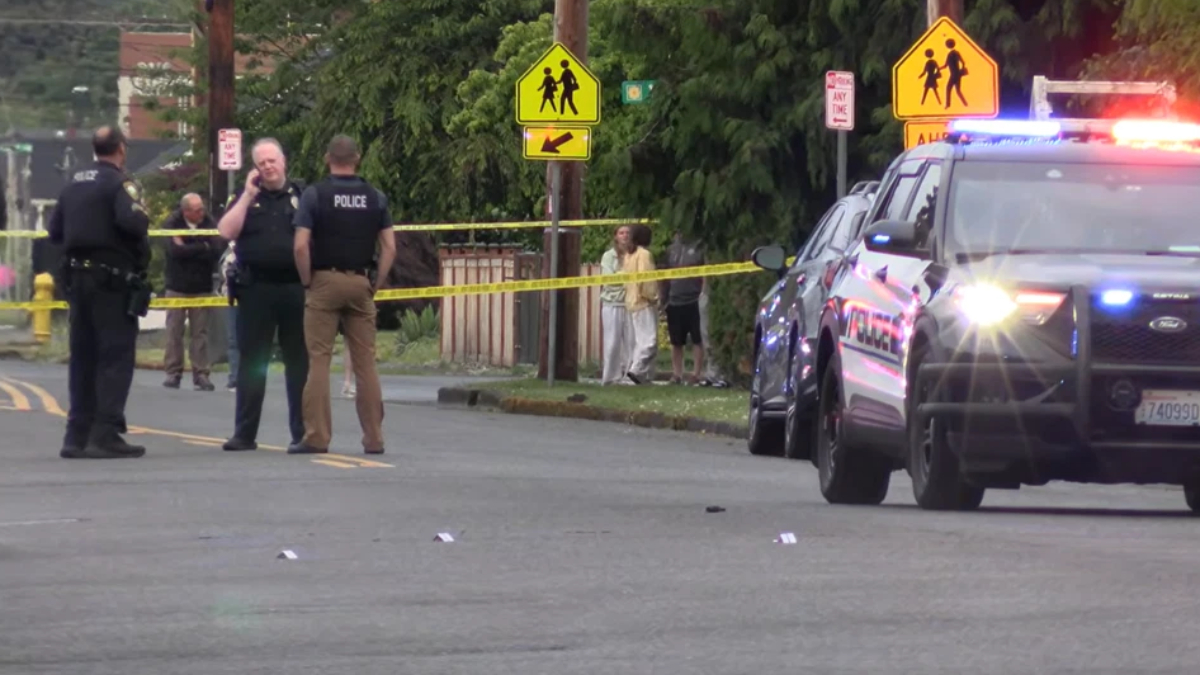 Police officers stand behind yellow crime-scene tape on a street, with flashing police car lights and bystanders in the background near a crosswalk sign.