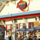 Sunny seaside diner with blue and purple striped umbrellas, outdoor tables, and patrons eating and chatting outside the bright white building.