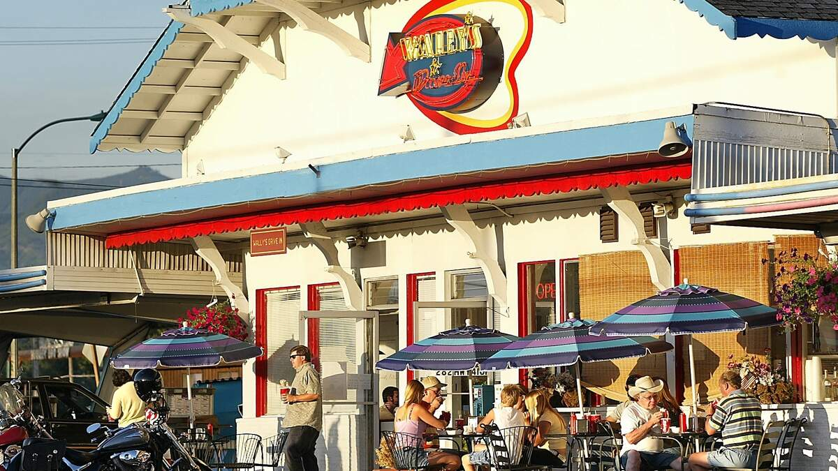 Sunny seaside diner with blue and purple striped umbrellas, outdoor tables, and patrons eating and chatting outside the bright white building.