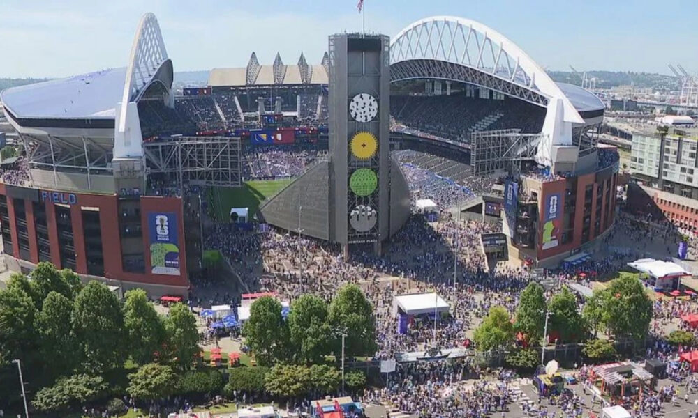 Aerial view of a large stadium filled with fans, with a tall central tower and surrounding concourses during daytime.