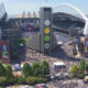Aerial view of a large stadium filled with fans, with a tall central tower and surrounding concourses during daytime.