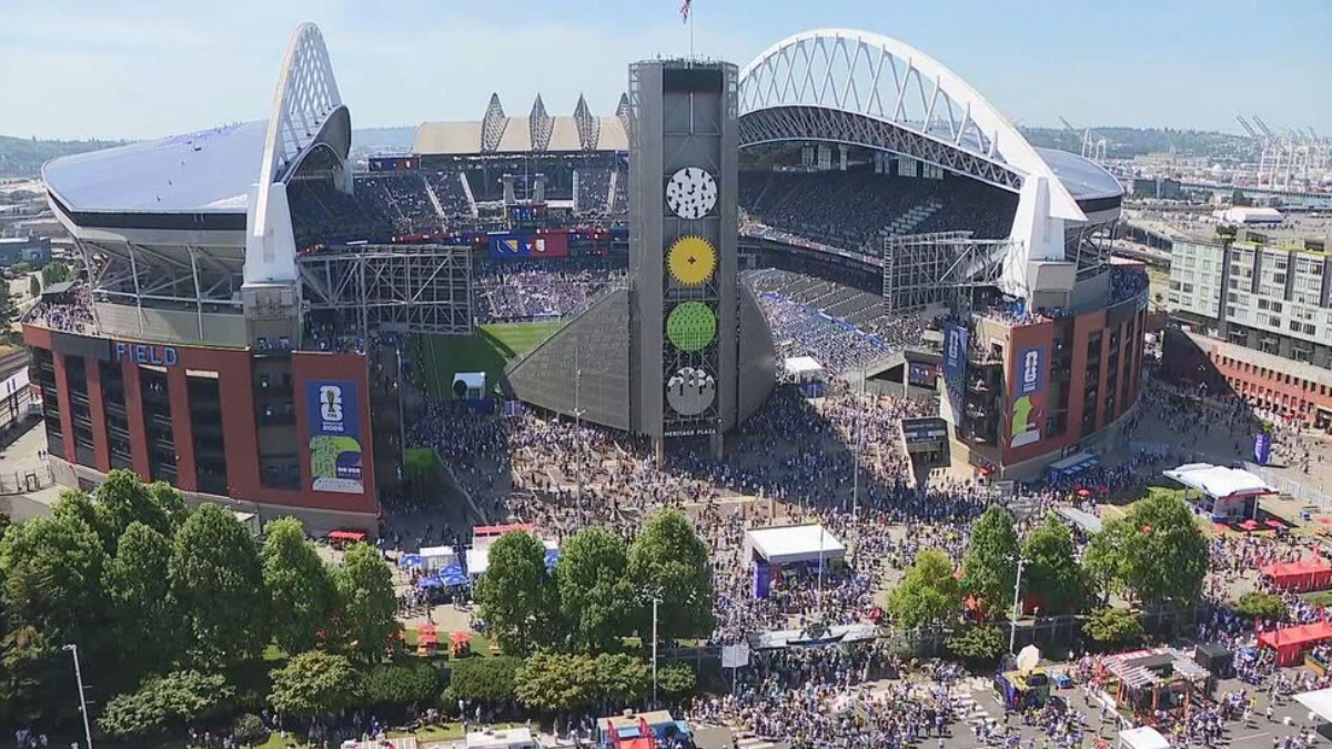 Aerial view of a large stadium filled with fans, with a tall central tower and surrounding concourses during daytime.