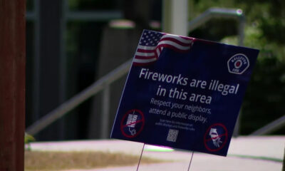 Blue sign on a lawn that reads, 'Fireworks are illegal in this area. Respect your neighbors, attend a public display,' with an American flag graphic in the corner.