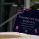 Blue sign on a lawn that reads, 'Fireworks are illegal in this area. Respect your neighbors, attend a public display,' with an American flag graphic in the corner.