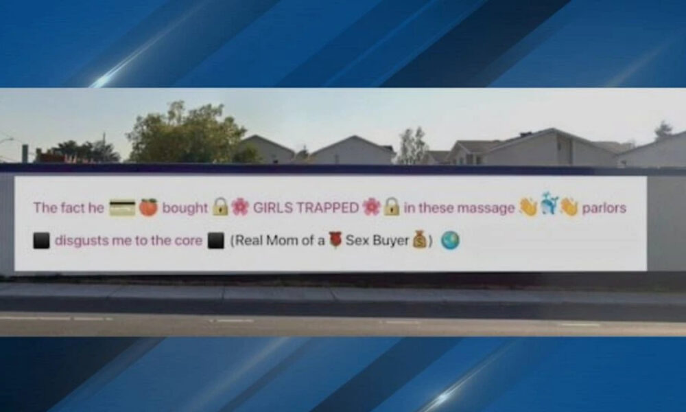 White rectangular banner on a fence with an anti-sex-trafficking message about girls trapped in massage parlors, plus emoji icons.