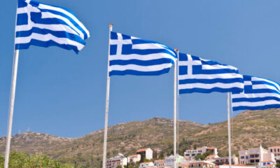 Four Greek flags fluttering on tall poles against a clear blue sky over a hillside town.