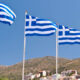 Four Greek flags fluttering on tall poles against a clear blue sky over a hillside town.