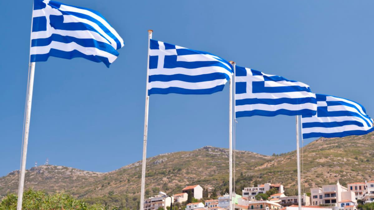 Four Greek flags fluttering on tall poles against a clear blue sky over a hillside town.