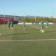 Soccer players in green jerseys practicing drills on a sunny artificial turf field with orange cones and a ball nearby.