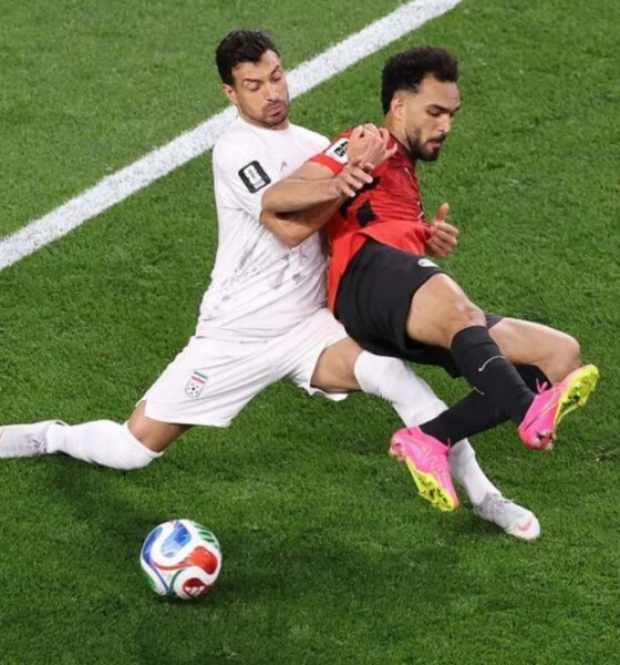 Two soccer players collide: a white-jersey defender tackles a red-and-black attacker near the sideline as the ball rests on the grass.