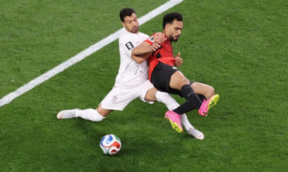 Two soccer players collide: a white-jersey defender tackles a red-and-black attacker near the sideline as the ball rests on the grass.