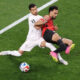 Two soccer players collide: a white-jersey defender tackles a red-and-black attacker near the sideline as the ball rests on the grass.