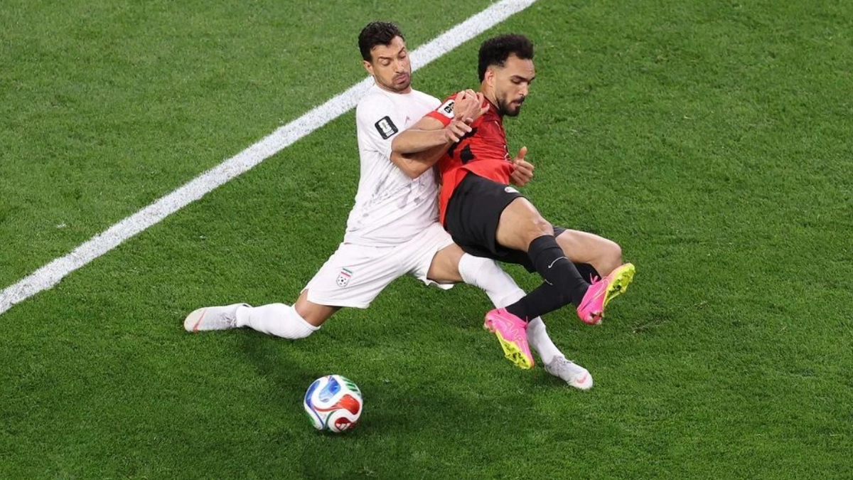Two soccer players collide: a white-jersey defender tackles a red-and-black attacker near the sideline as the ball rests on the grass.