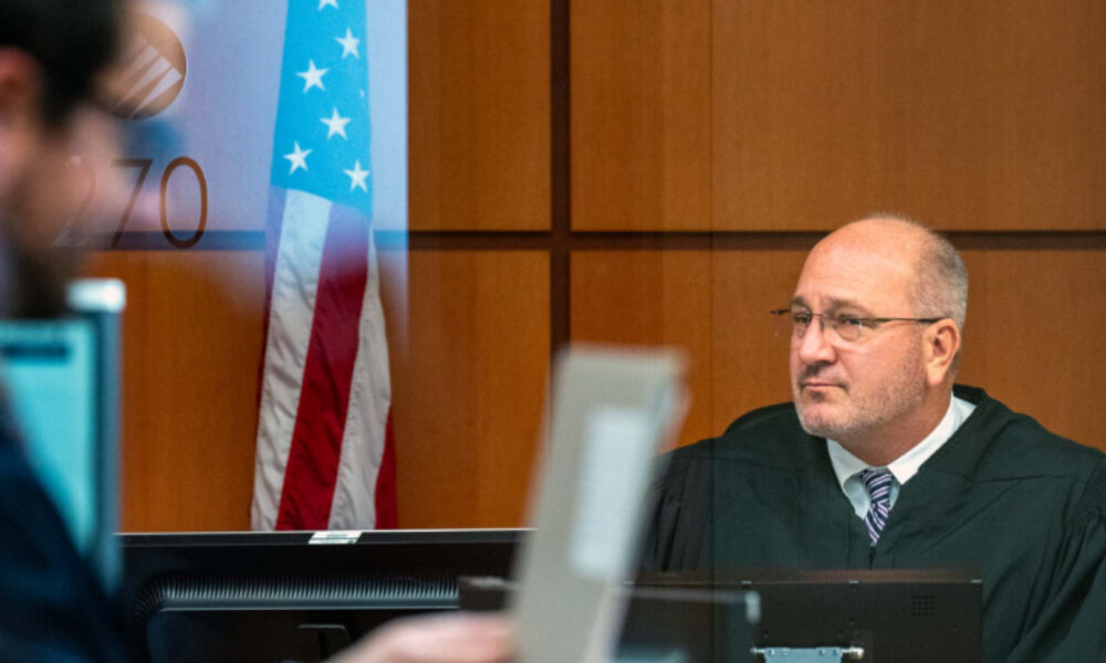 A judge in a black robe sits at a courtroom bench, with an American flag in the background.
